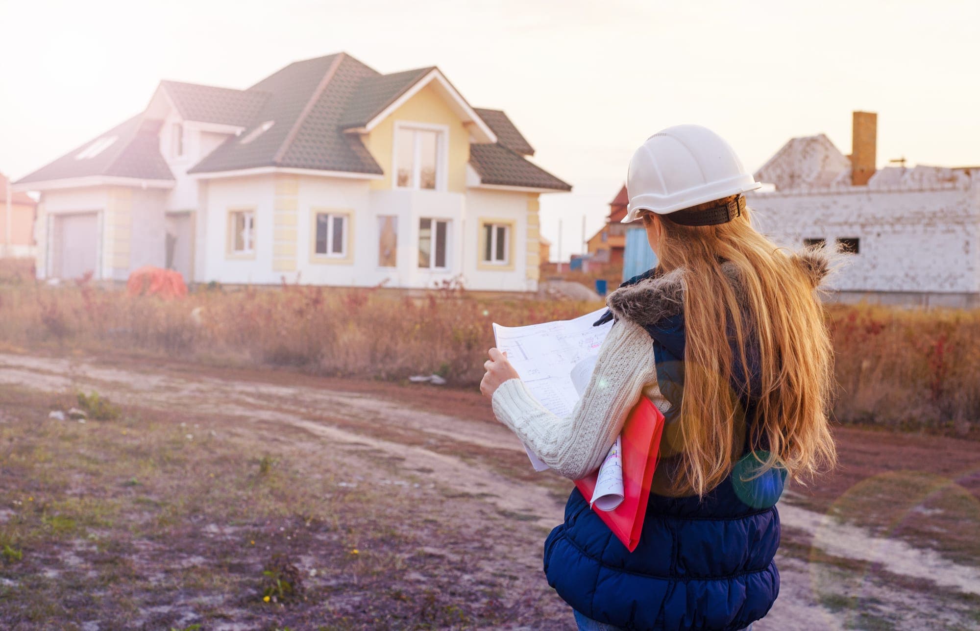 Builder in a hard hat reviewing plans in front of homes under construction
