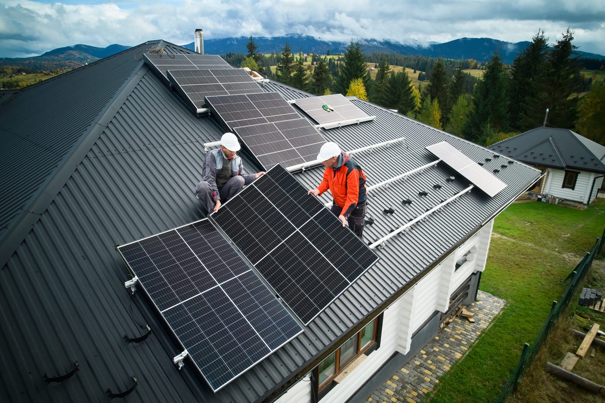 Two installers fitting solar panels on a metal roof with mountains behind